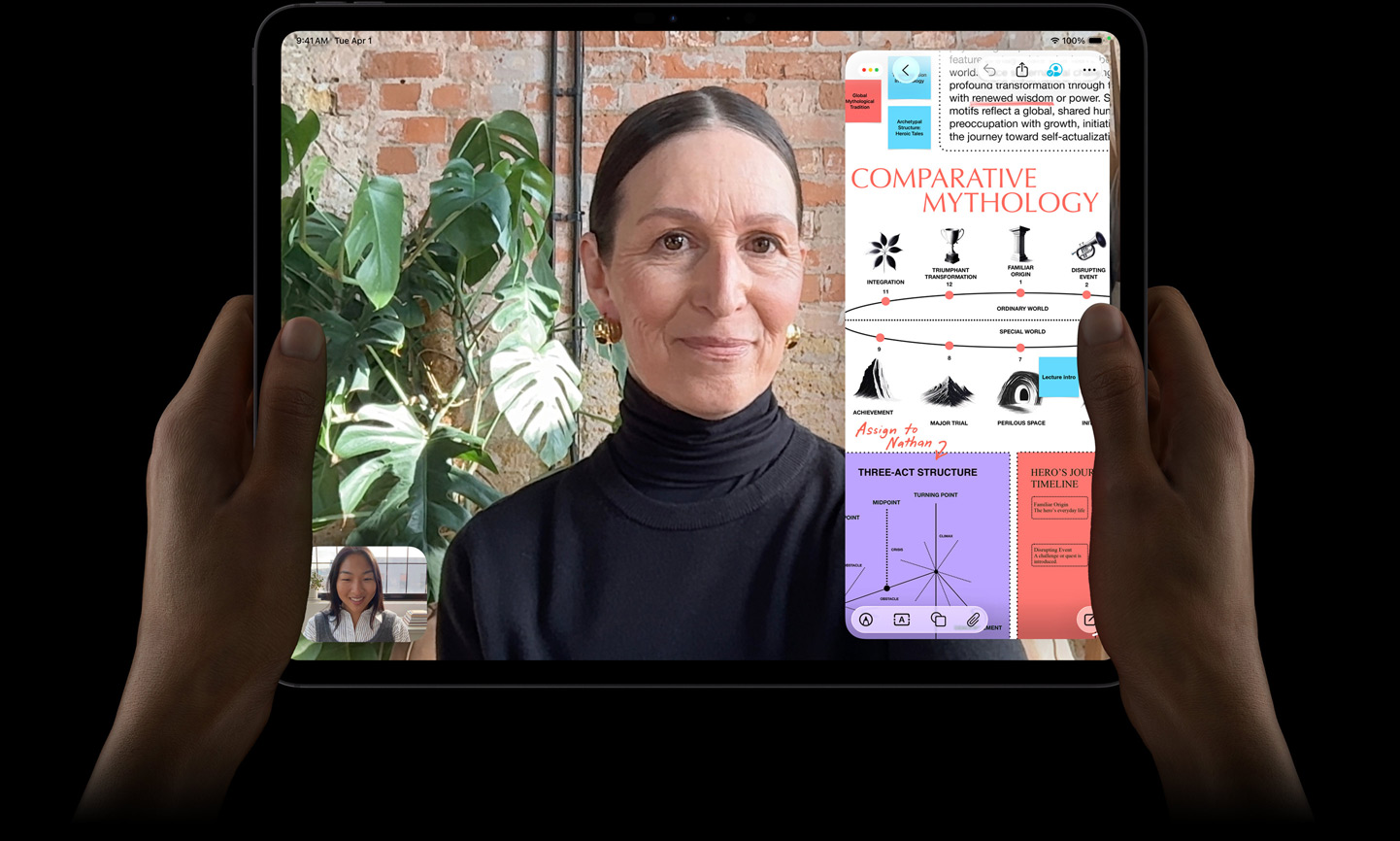 iPad Pro i space black vist forfra, et par hænder holder enheden i vandret position, afrundede hjørner, sort skærmkant; skærmen viser en FaceTime-samtale, der oversættes live, og en uddannelsesapp, der viser en præsentation af “Comparative Mythology” med tekst, diagrammer og håndskrevne noter