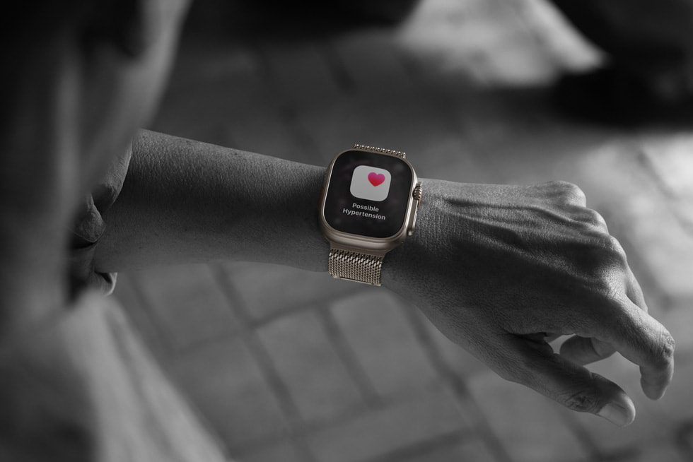 Une photo en noir et blanc d’une personne regardant son Apple Watch Ultra 3 qui affiche une notification d’hypertension accompagnée d’une icône rouge en forme de cœur.