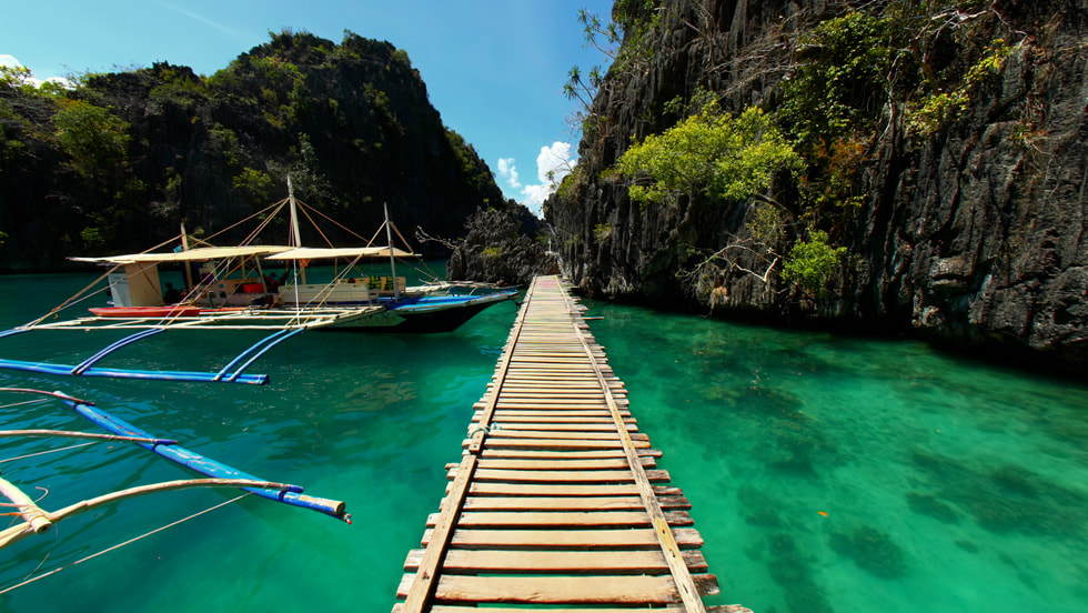 Imagen tomada en un Apple Vision Pro que muestra un paisaje de Explore POV en el que se ve un camino de tablas de madera que se extiende a la vera de una costa con aguas cristalinas. A la izquierda, se ve un bote de diseño tradicional que flota en el agua. 