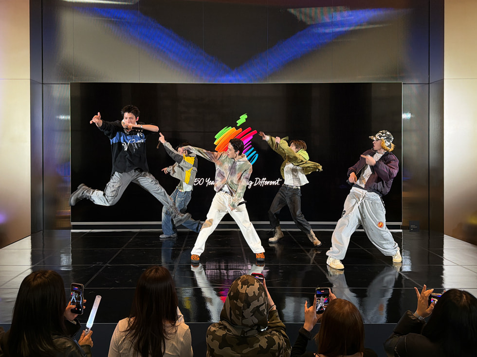 Members of the band CORTIS are pictured dancing on a small stage, with one member captured jumping in mid-air, at Apple Myeongdong in Seoul.