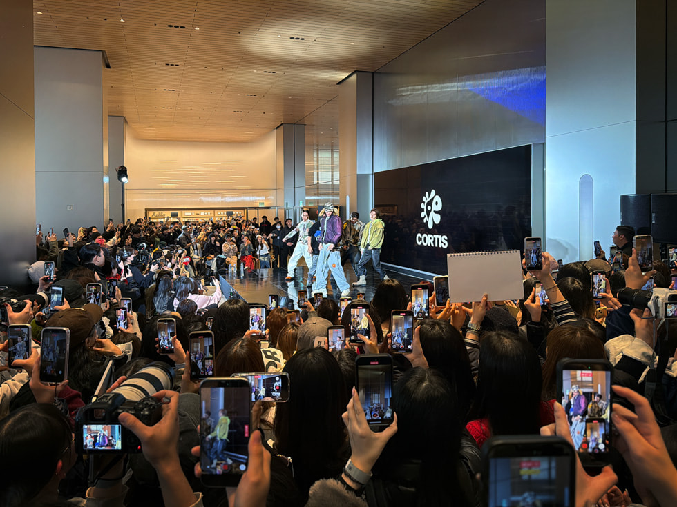 A view from the audience shows fans holding cameras and iPhone devices as CORTIS performs on a small stage at Apple Myeongdong in Seoul.