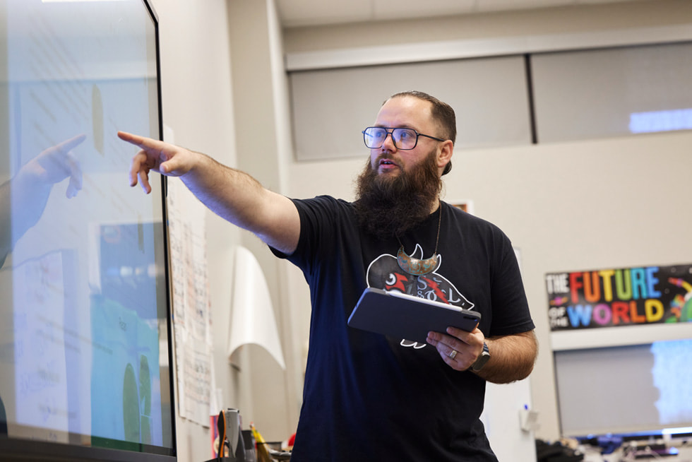 Tyler Teague sosteniendo un iPad y apuntando a una pantalla en un aula.