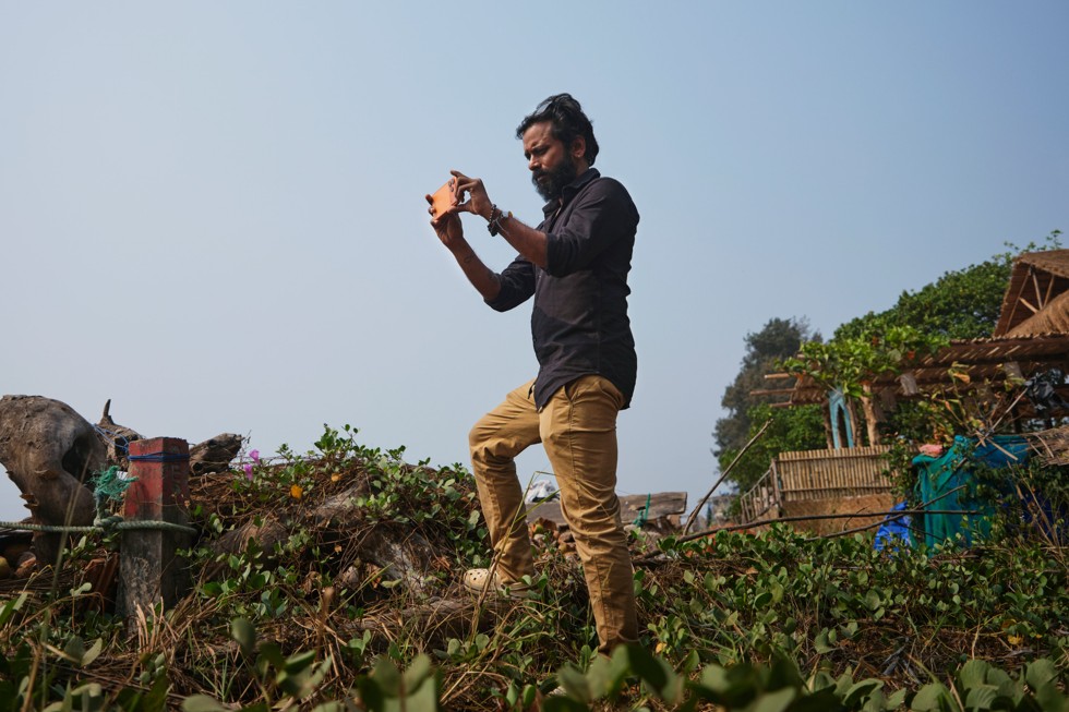 Ritesh Sharma stands on a grassy hill overlooking water, holding iPhone 17 Pro Max while filming.