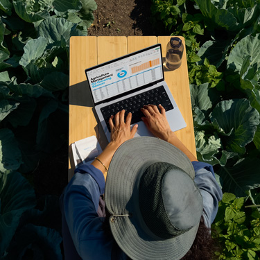 Vista panorámica de una mujer con un sombrero de ala ancha que trabaja en una presentación de negocios en un MacBook, sentada en una mesa en un jardín.