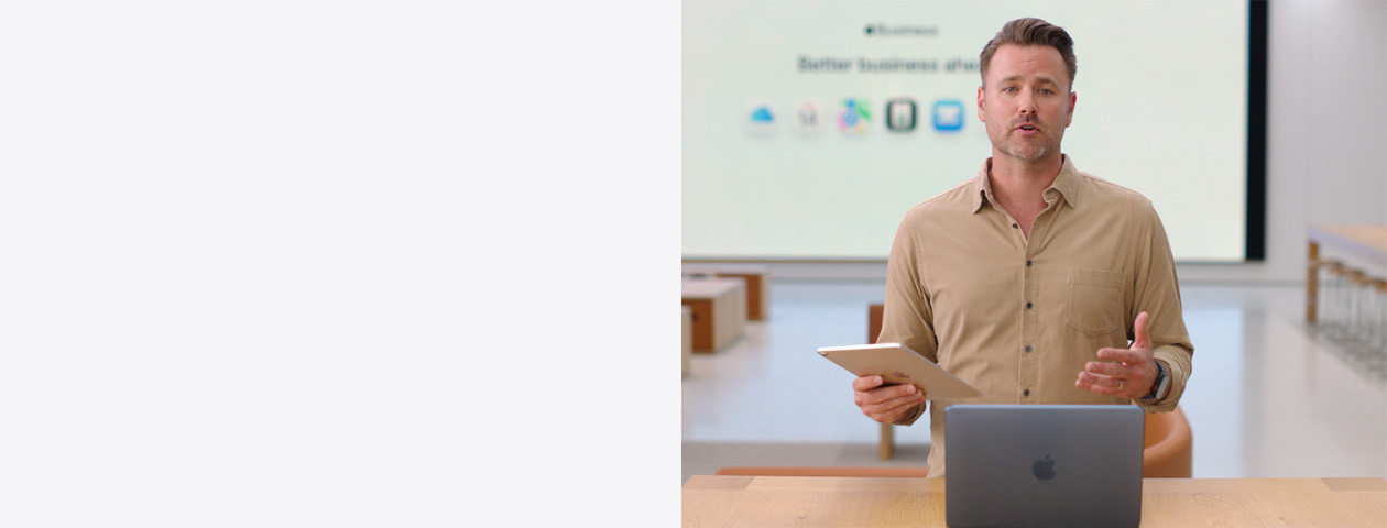 Présentateur parlant avec un iPad à la main, debout devant un MacBook posé sur une table, tandis qu’un grand écran derrière lui affiche les icônes de plusieurs apps professionnelles