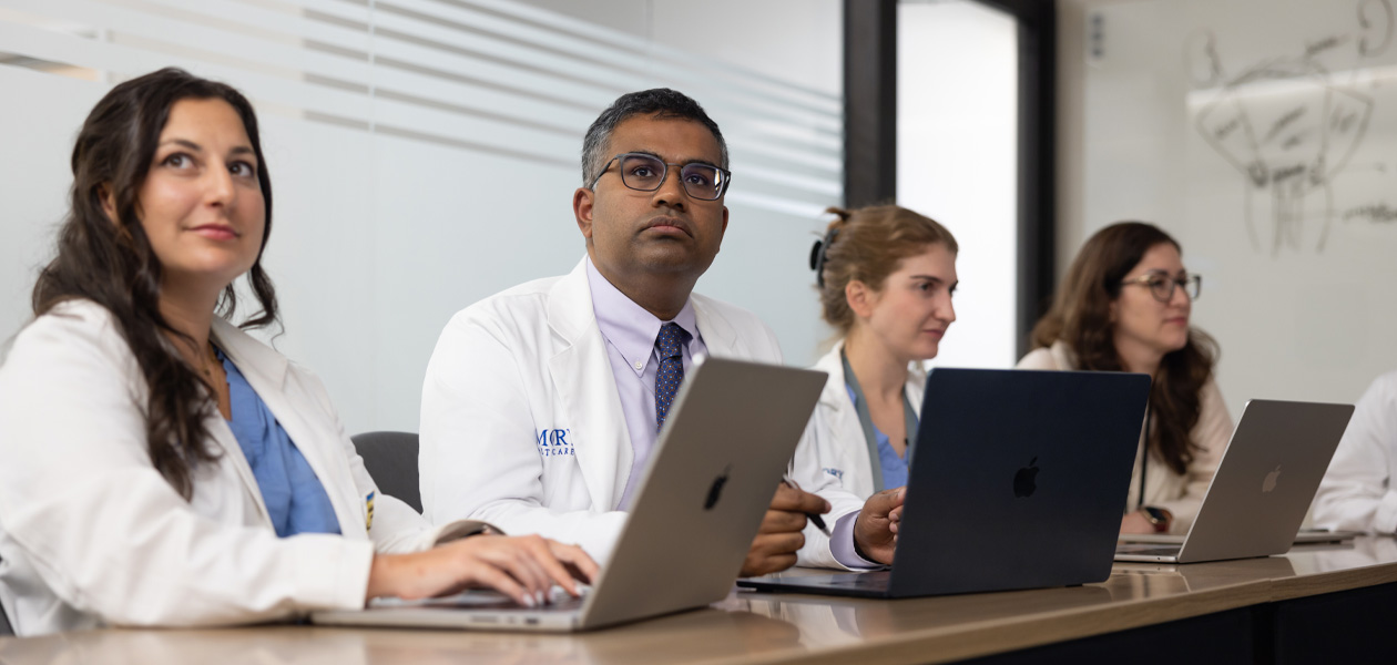 Four colleagues in doctors’ coats sit in a row, each working on a MacBook.