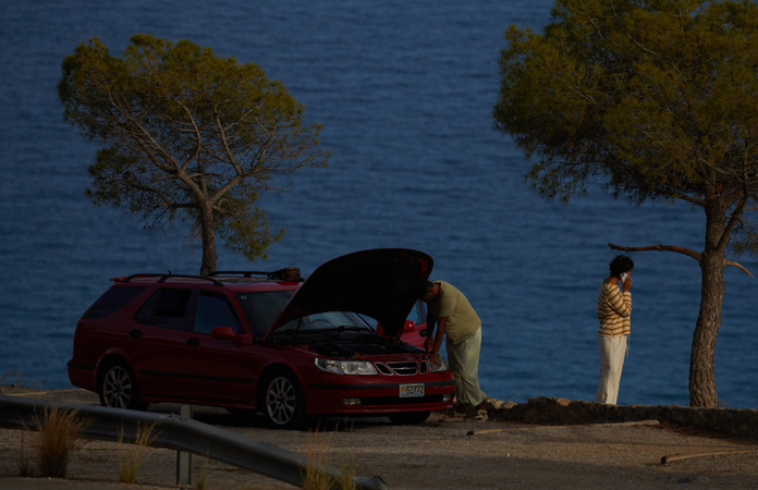 A photo of a car stranded by the side of a road, one person by the car using Roadside Assistance via satellite on iPhone