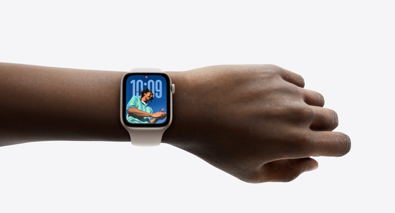 Pohled zblízka na stříbrné Apple Watch s gumovým řemínkem nasazené na zápěstí, je vidět aplikace Hodiny v novém Liquid Glass designu